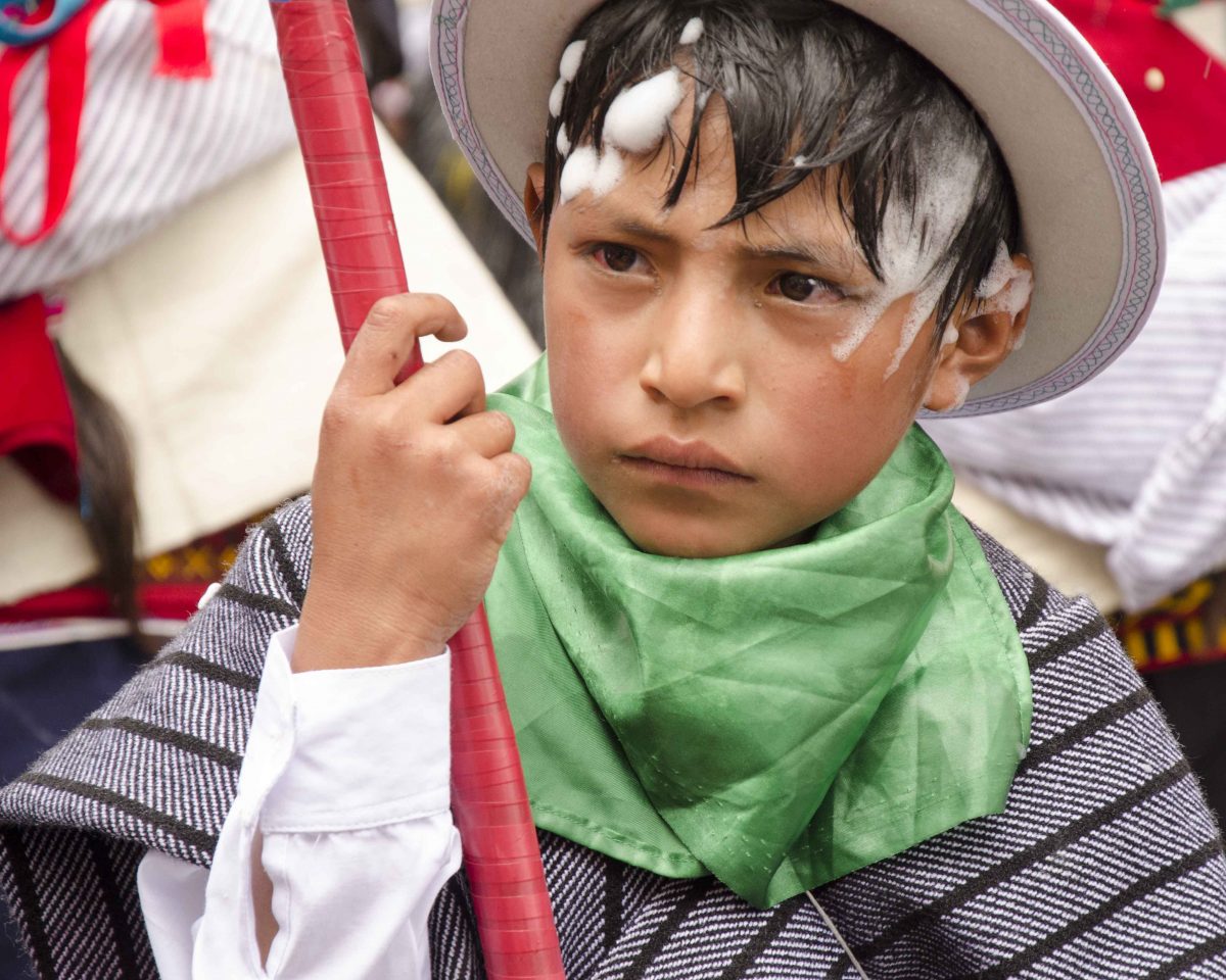 A young boy walks the parade route; Carnival in Guamote, Ecuador | ©Angela Drake Young boy with serious face is dressed in gray poncho and white wool hate