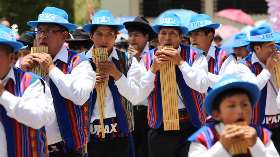 Musicians dressed in white shirts, rainbow color vests, and blue sky hats play wind instruments
