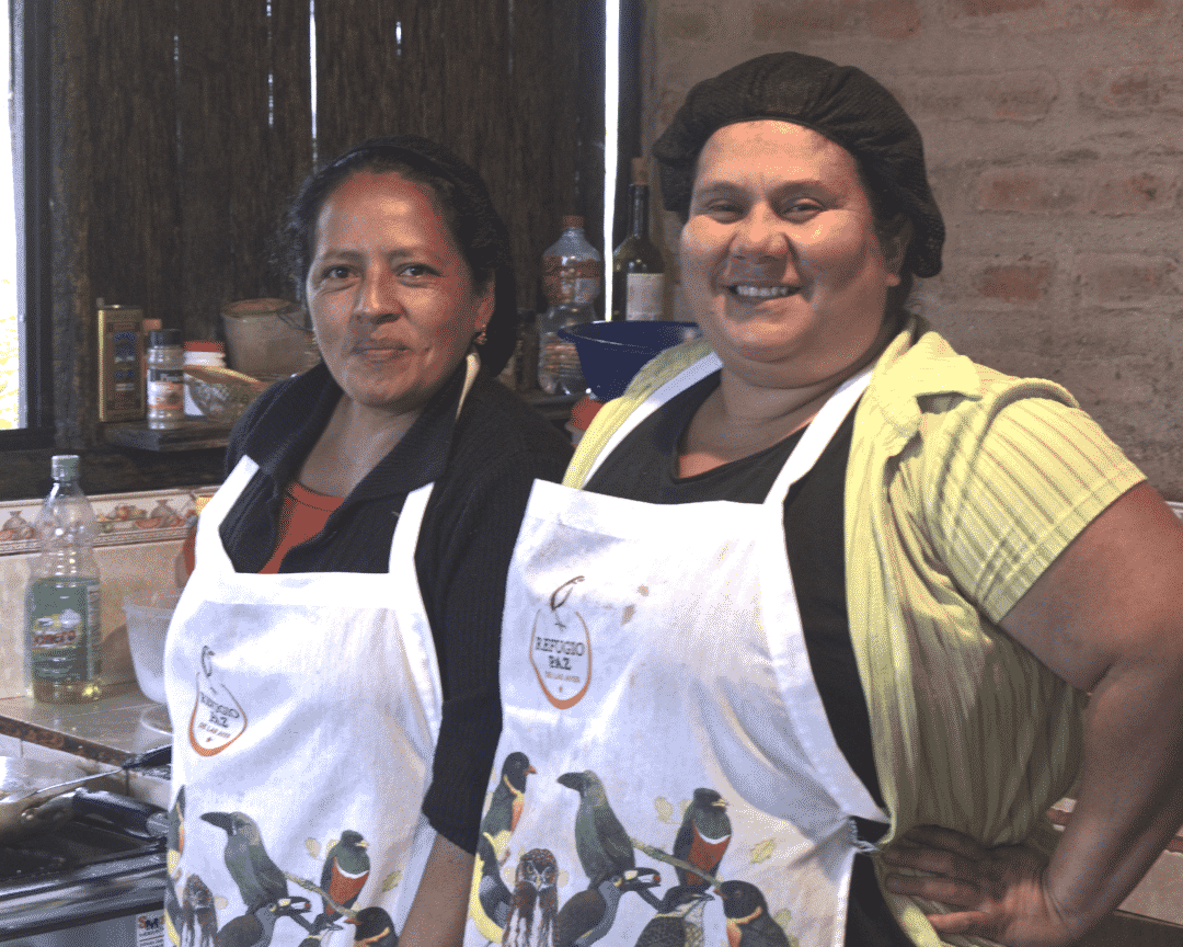 Two smiling women, wearing aprons with many pictures of birds, stand smiling in a kitchen