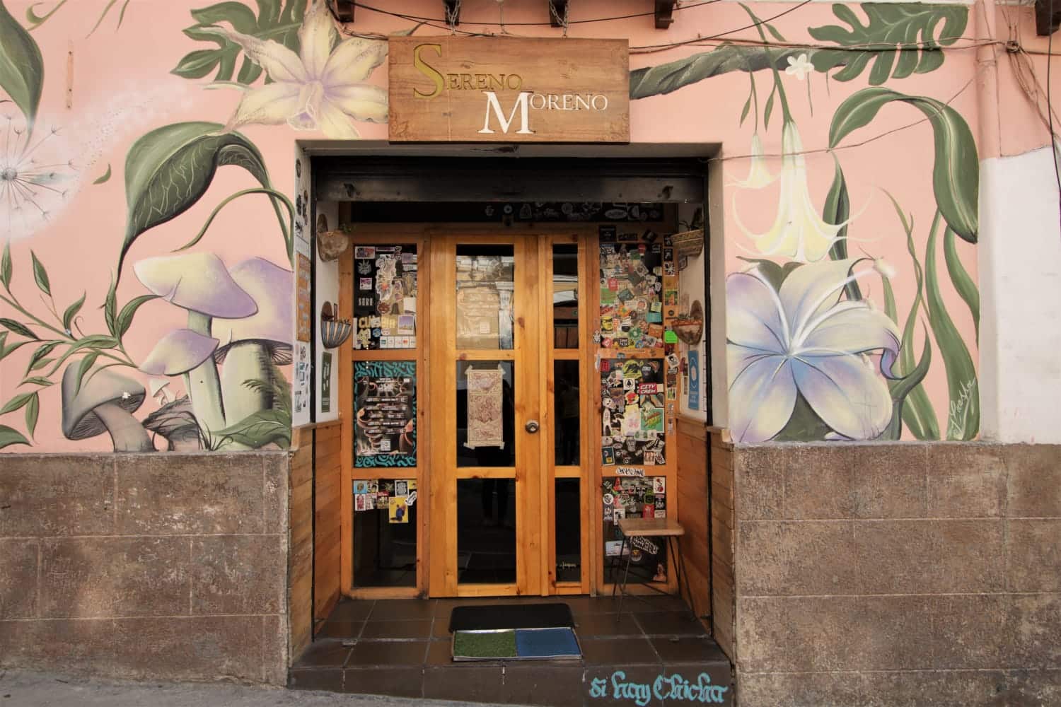 Front door of the Sereno Moreno Picantería, La Tola, Quito, Ecuador | ©Sereno Moreno Picantería Front door of Sereno Moreno is made of wood and glass. The front wall is painted with a mural of flowers white flowers, green leaves and stems, with a pale pink background.