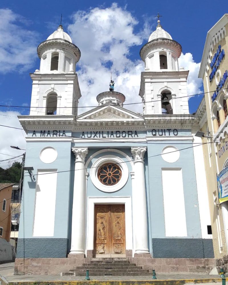 An old church has a wooden door, a rose window, two white towers, and walls painted light blue with white accents.