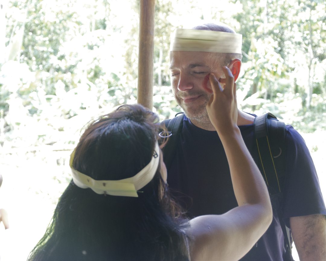 A tall, white, male tourist is having his face painted with red markings by a Huaorani woman