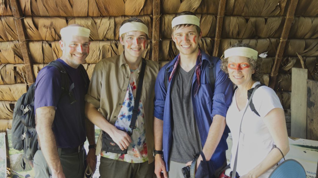 A US American, white family of four poses with face paint made of achiote seeds. A tall man stands to the left, two teenage boys in the middle, and a short woman stands on the right.