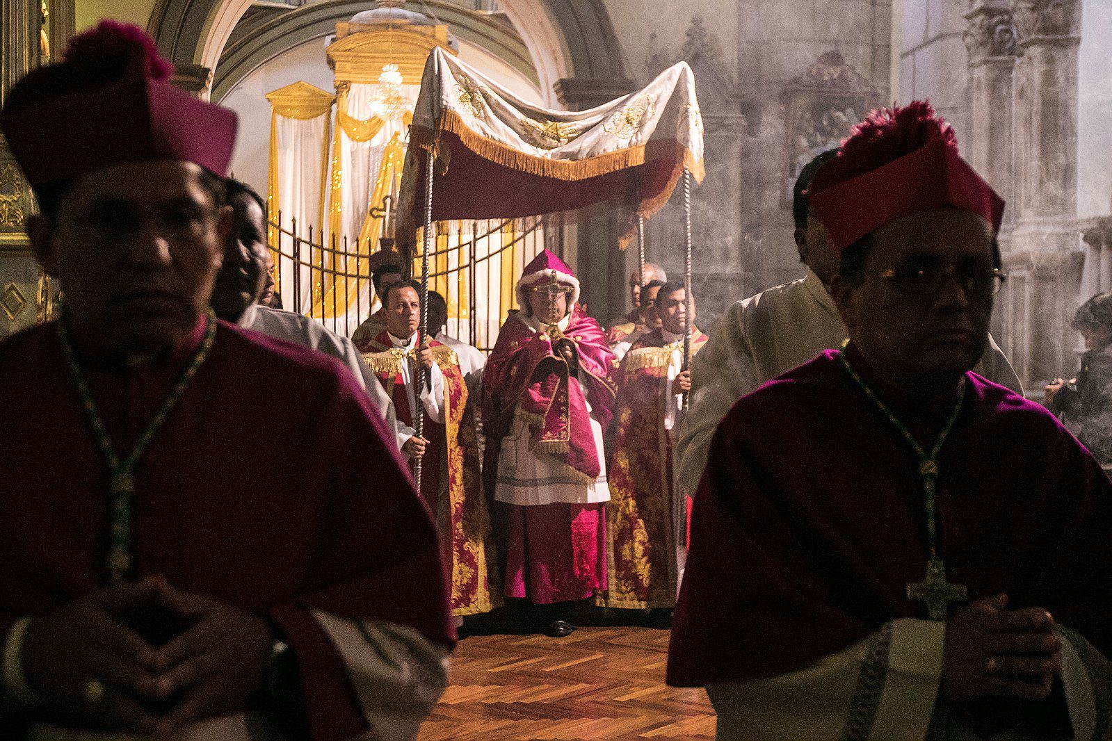 A hooded priest holding a golden cross before him stands beneath a canopy help by men dressed in robes.