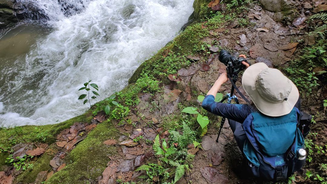 Woman crouches down to a camera tripod as she frames a picture of a waterfall