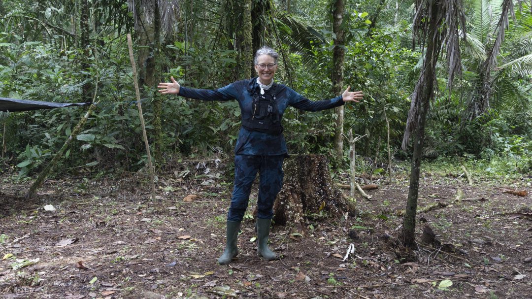 Angie stands with arms spread wide to show her long sleeved shirt and long pants tucked into rubber boots, suggested measures to prevent mosquito bites in the Amazon