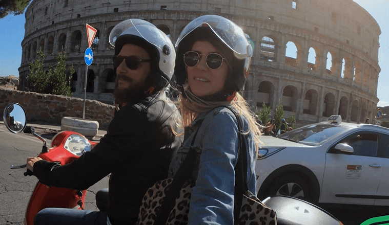 A man and woman take a selfie while sitting on a moped in front of the Coliseum in Rome