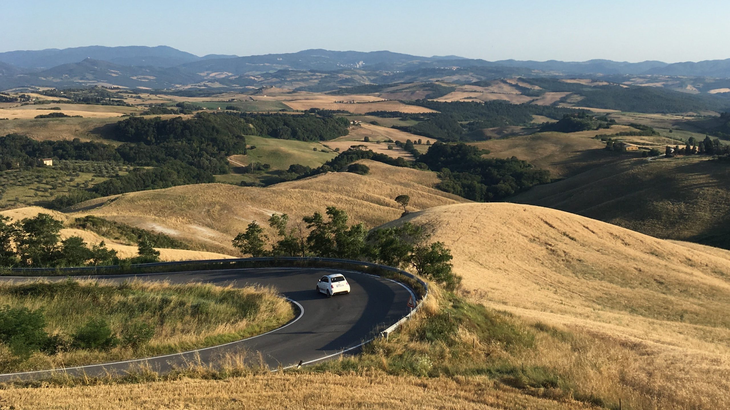 A small white car driving in Italy takes a sharp turn on a paved road with views of the stunning Italian countryside.