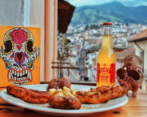 Foreground: delicious plate of food, bottle of chicha, and Ecuadorian art; background: view of Quito