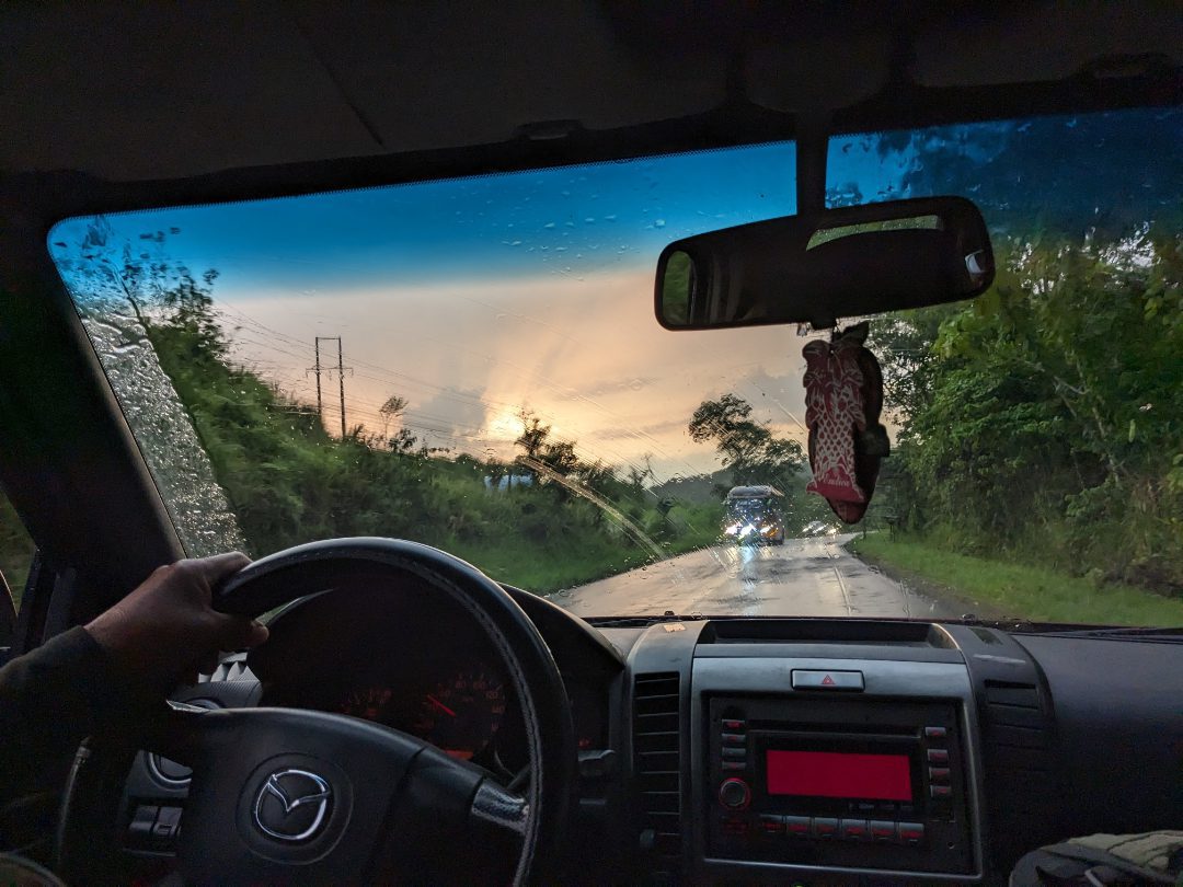 A view of a rainy dawn on a highway with oncoming traffic from the backseat looking out the front window past the driver's wheel of an SUV