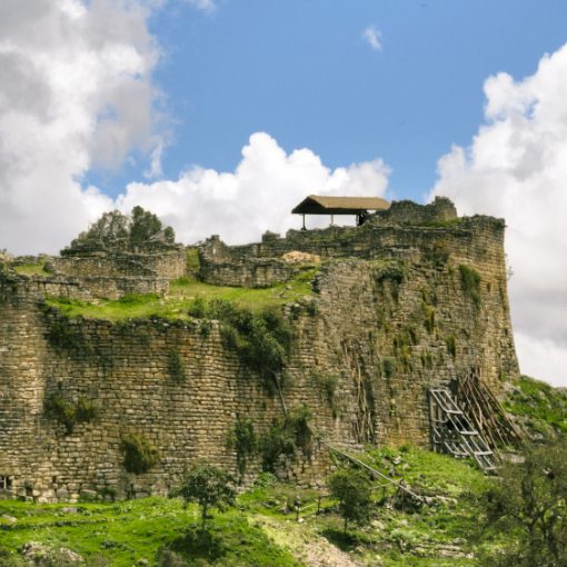 View of Kuelap’s massive stone walls rising dramatically from the mountaintop, set against a backdrop of a cloudy blue sky.