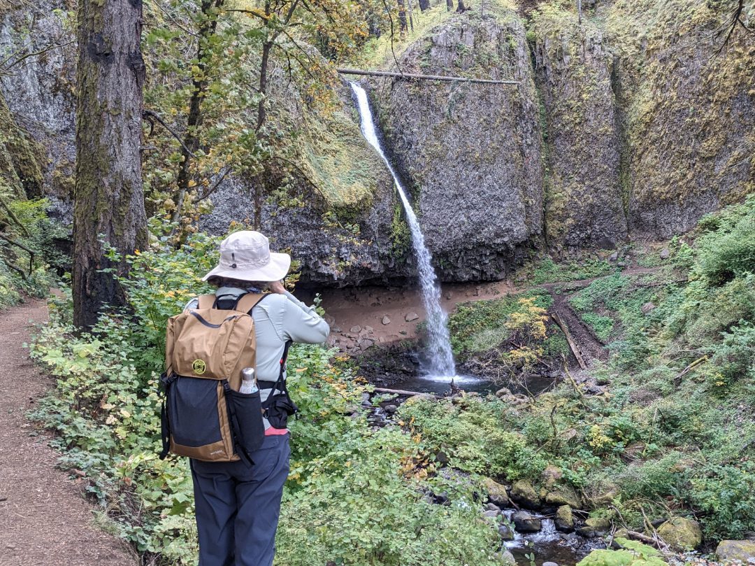 A woman photographing a waterfall while carrying the Wy'east Rucksack