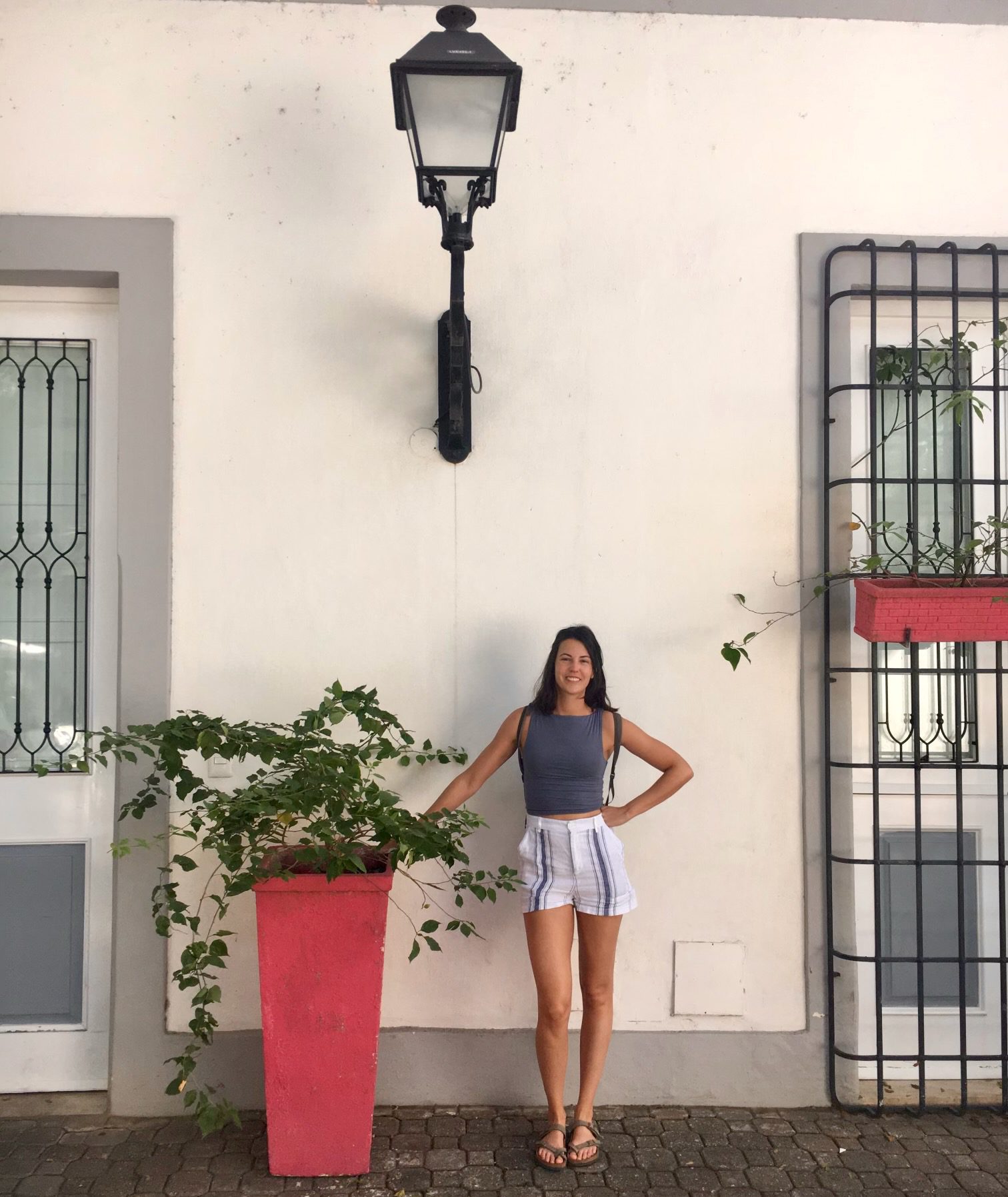 A woman in shorts and tank top poses before a white wall next to a tall planter