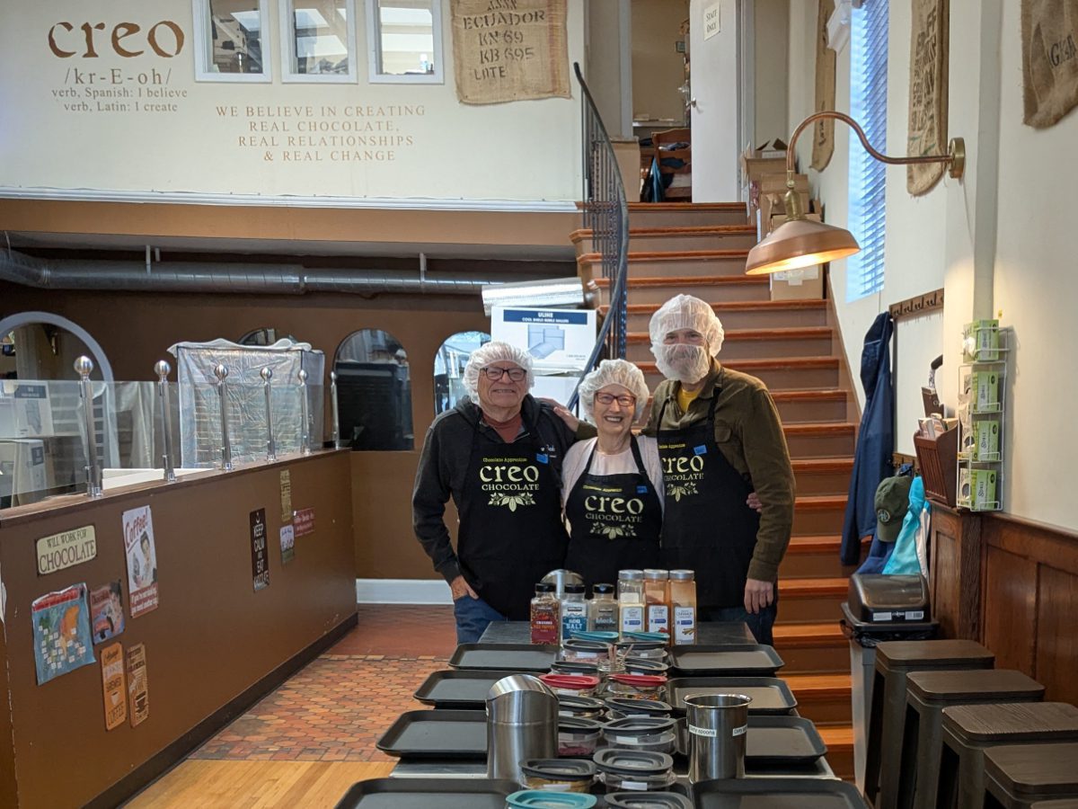 Three people stand at the end of a long prep table, dressed in Creo apprentice aprons, hair nets and  a beard net for one gentleman. 