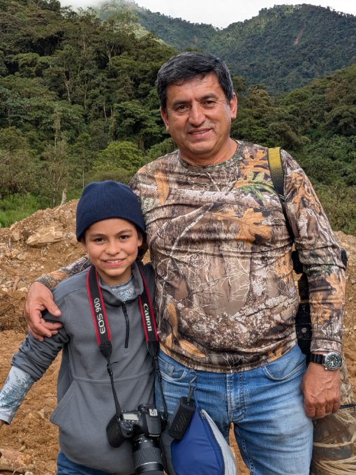 A father poses with his arm around his young son with hills and forest in the background