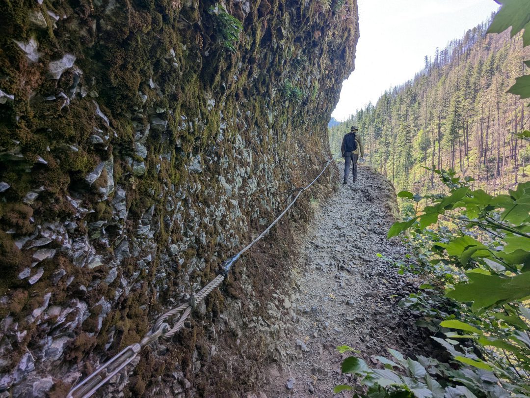 A lone hiker climbs a narrow trail on the edge of a cliff, cables along the mountainside to the left, a drop off and valley view to the right