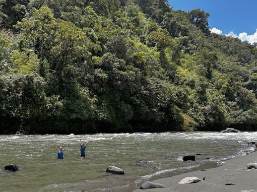 Two women stand in the Rio Quijos river, arms raised in joy of cold water immersion
