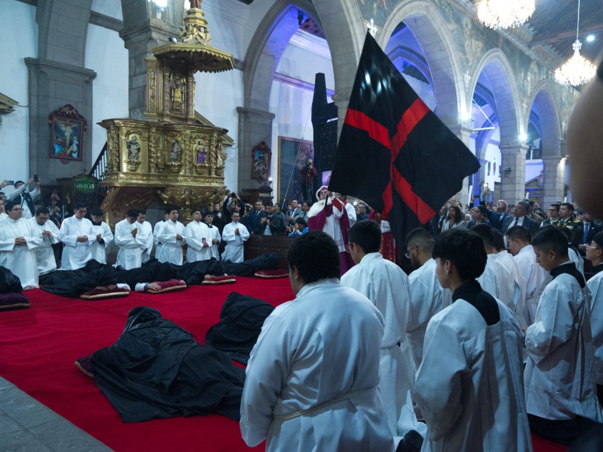 Archbishop waving ceremonial flag over prone black-robed priests as white-robed clergy look on, Arrastre de Caudas Quito
