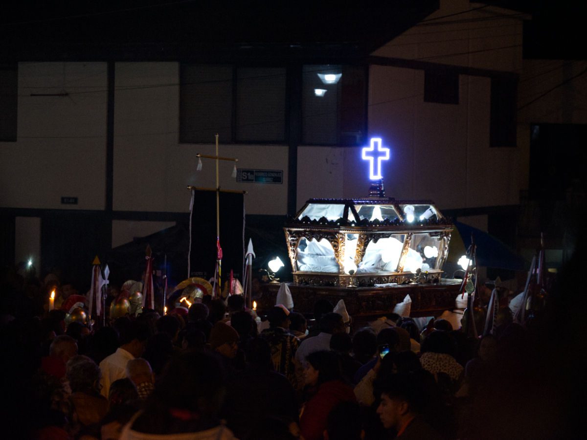 A clear glass casket with the wrapped wooden body of Christ is held aloft by several men dressed in white and topped with a neon-lit, purple cross.