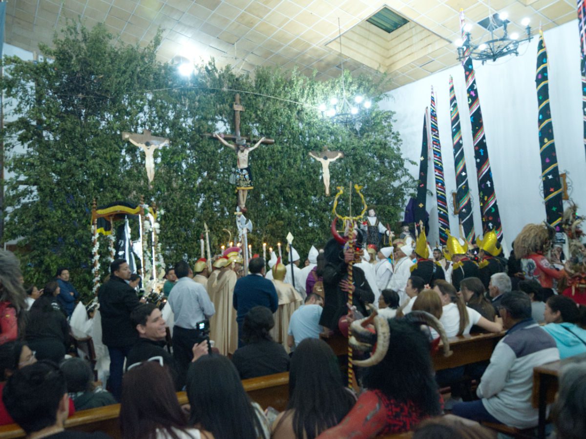People sitting in pews, characters from the reenactment of the Good Friday fill the front of the church with a backdrop of Jesus on the Cross and the Almas Santas placed along the right wall.